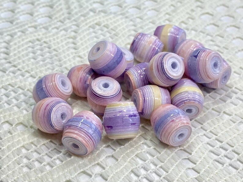 K&ouml;nnte beinhalten: Eine Ansammlung von tonnenf&ouml;rmigen Perlen mit einem geschichteten, gerollten Papierdesign. Jede Perle zeigt eine Farbpalette aus zarten pastellfarbenen violetten, hellrosa, cremefarbenen und wei&szlig;en Streifen mit einem mittigen Loch.