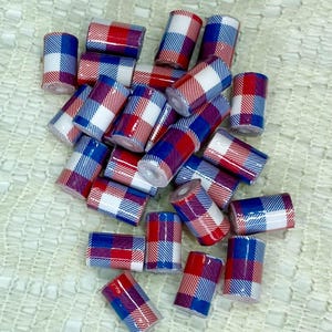 May include: A collection of small, cylindrical beads with a plaid pattern in red, white, and blue. The beads are scattered across a textured, off-white surface, creating a visually appealing arrangement. The ends of the beads are visible, showing a hollow center.