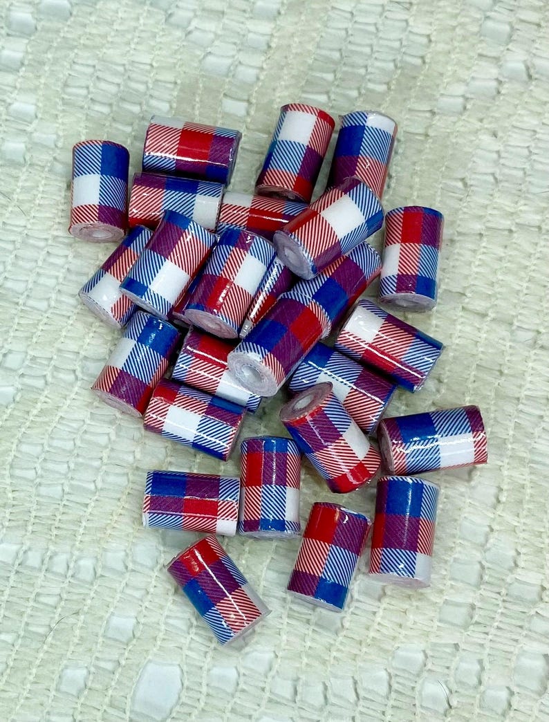 May include: A collection of small, cylindrical beads with a plaid pattern in red, white, and blue. The beads are scattered across a textured, off-white surface, creating a visually appealing arrangement. The ends of the beads are visible, showing a hollow center.