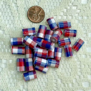 May include: A collection of small, cylindrical beads with a red, white, and blue plaid pattern. A U.S. penny is included for size reference. The beads are scattered on a textured, off-white surface.
