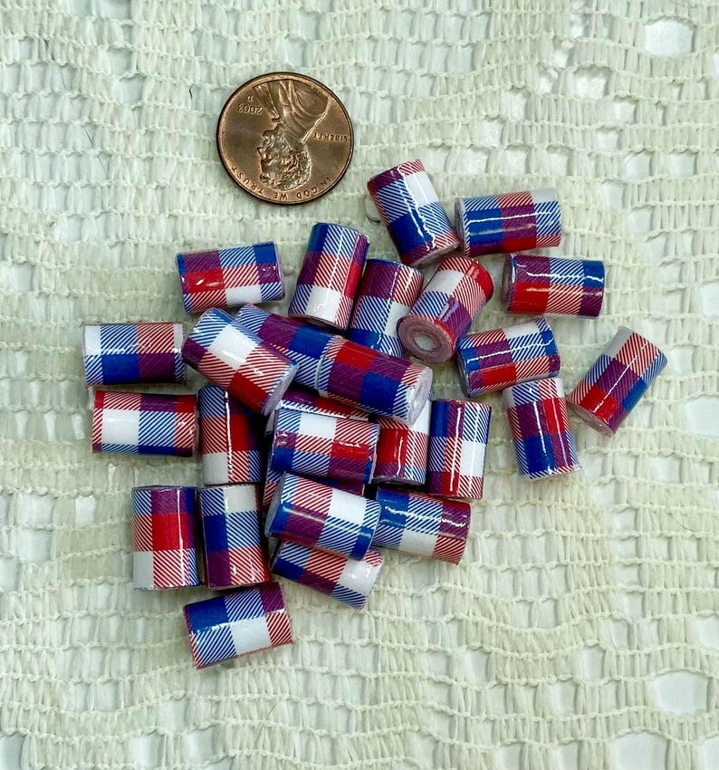 May include: A collection of small, cylindrical beads with a red, white, and blue plaid pattern. A U.S. penny is included for size reference. The beads are scattered on a textured, off-white surface.