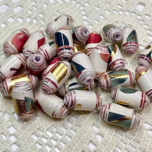 May include: A collection of cylindrical paper beads featuring a white base with assorted patterns in red, dark teal, and metallic gold. Each bead has a central hole and a smooth, glossy finish, displaying layered paper construction with visible edges.