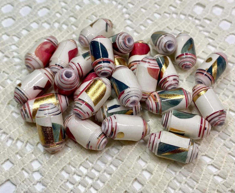 May include: A collection of cylindrical paper beads featuring a white base with assorted patterns in red, dark teal, and metallic gold. Each bead has a central hole and a smooth, glossy finish, displaying layered paper construction with visible edges.