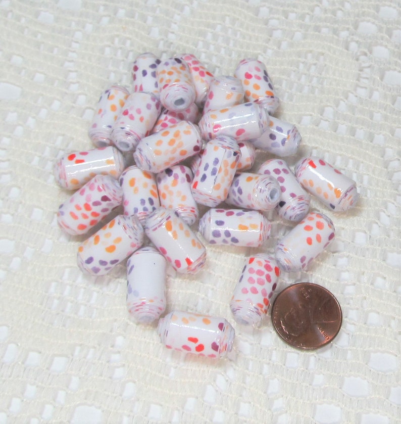May include: A pile of white paper beads with orange, pink and purple polka dots. The beads are approximately 2.5 cm long and are arranged in a random pattern. A penny is included for scale.