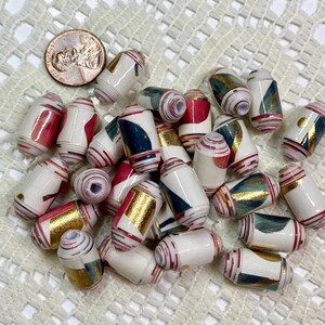 May include: A collection of cylindrical paper beads featuring a white base with abstract geometric patterns in red, dark blue, and metallic gold foil accents. A copper-colored penny is included for scale to show the size of the individual beads.