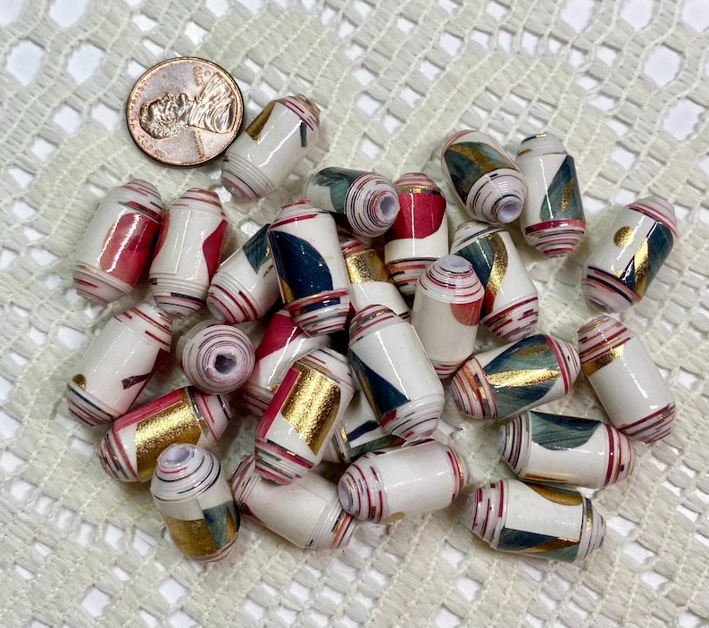 May include: A collection of cylindrical paper beads featuring a white base with abstract geometric patterns in red, dark blue, and metallic gold foil accents. A copper-colored penny is included for scale to show the size of the individual beads.