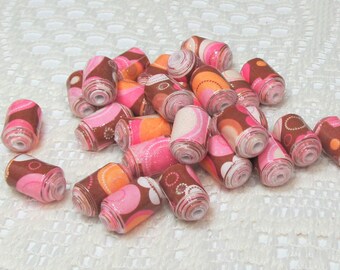 May include: A collection of paper beads in shades of pink, brown, and orange. The cylindrical beads have a patterned design and are scattered on a white, textured surface. These beads are suitable for jewelry making and other craft projects.