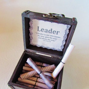 May include: A wooden box with a hinged lid and a metal clasp. The box is open and contains several rolled up pieces of paper. The inside of the lid has a quote about leadership: "A leader is one who knows the way, goes the way, and shows the way." - John Maxwell