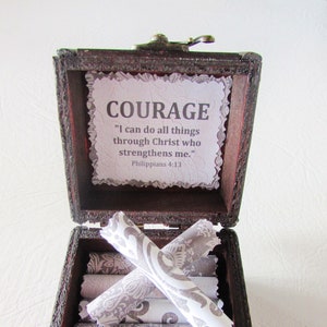 May include: A wooden box with a hinged lid and a metal latch. The box is open and contains several rolled up pieces of paper with a gray and white pattern. The inside of the box has a paper insert with the words "COURAGE" and the quote "I can do all things through Christ who strengthens me." Philippians 4:13.