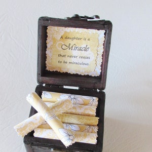 May include: A wooden box with a quote on the lid that reads "A daughter is a miracle that never ceases to be miraculous." The box is open and contains several rolled up pieces of paper with yellow and white floral patterns.