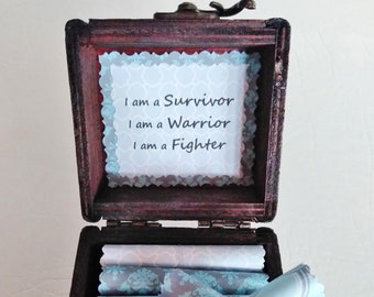 May include: A wooden box with a hinged lid, open to reveal a stack of rolled up blue and white patterned paper. The inside of the lid has a white card with the text "I am a Survivor, I am a Warrior, I am a Fighter."
