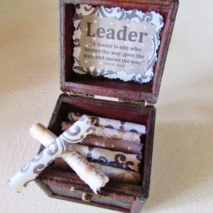 May include: A small, open wooden box with a dark red finish, containing rolled-up scrolls. The box has a decorative paper inside with the word "Leader" and a quote by John Maxwell. The scrolls are various colors and patterns.