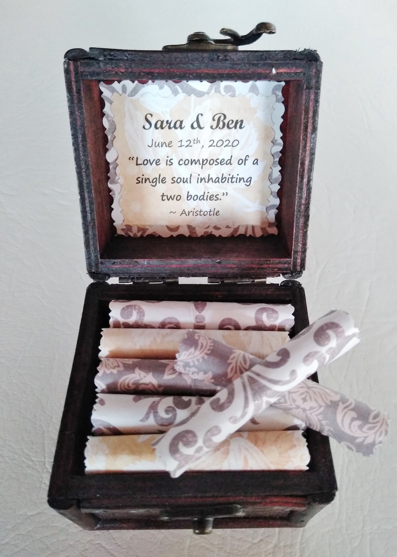 Pictured is an open wood box. Inside are rolled scrolls, each have a love quote. On the inside lid on decorative paper are the couple's names, wedding date, and quote, Love is composed of a single soul inhabiting two bodies, Aristotle.