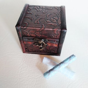 May include: A small, brown wooden box with a floral embossed design and a metal latch. The box is sitting on a white surface with two rolled up pieces of fabric with a floral pattern.