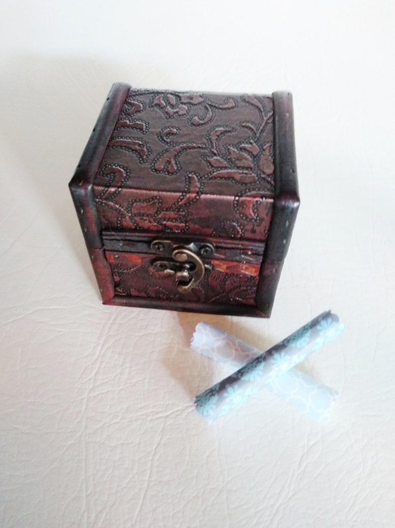 May include: A small, brown wooden box with a floral embossed design and a metal latch. The box is sitting on a white surface with two rolled up pieces of fabric with a floral pattern.