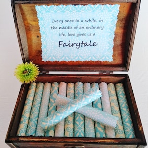 May include: A wooden box with a blue and white patterned paper lining. The box is open and contains several rolled up pieces of paper with the same pattern. The lid of the box has a quote that reads: "Every once in a while, in the middle of an ordinary life, love gives us a Fairytale."