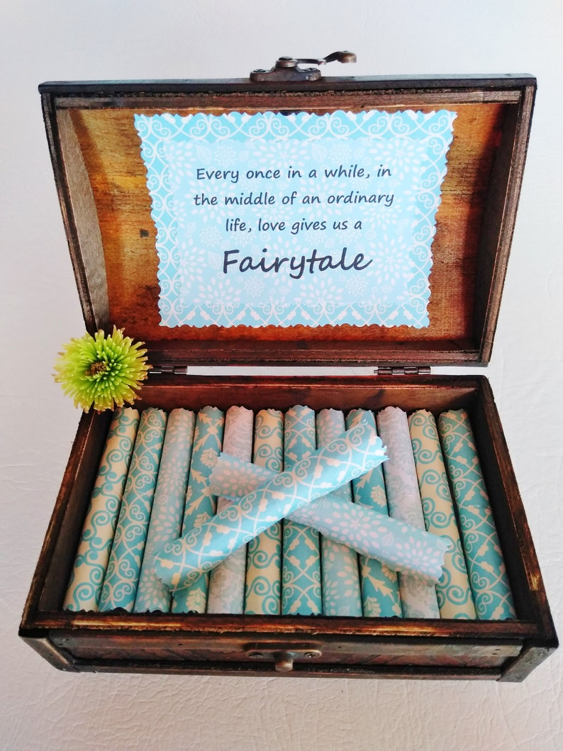 May include: A wooden box with a blue and white patterned paper lining. The box is open and contains several rolled up pieces of paper with the same pattern. The lid of the box has a quote that reads: "Every once in a while, in the middle of an ordinary life, love gives us a Fairytale."