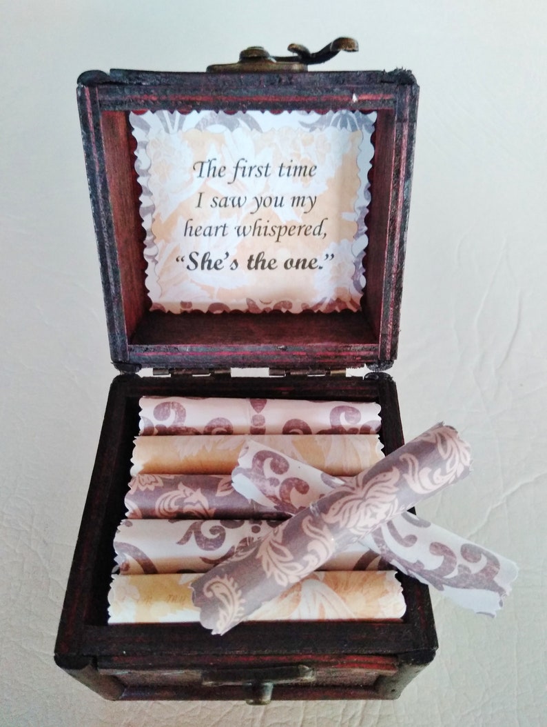 May include: A wooden box with a hinged lid, open to reveal a stack of rolled paper scrolls. The inside of the lid has a printed message that reads "The first time I saw you my heart whispered, "She's the one.""