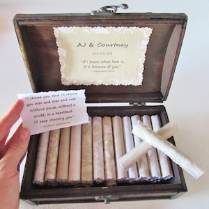 May include: A wooden box with a hinged lid, open to reveal a collection of rolled up papers inside. The box is decorated with a small card that reads "AJ & Courtney 10/13/13" and a quote by Hermann Hesse. The card is decorated with a floral pattern. The rolled up papers are also decorated with a floral pattern.