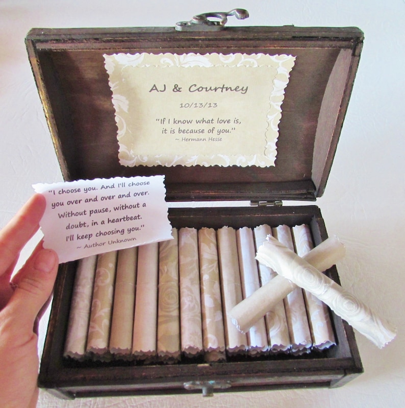 May include: A wooden box with a hinged lid, open to reveal a collection of rolled up papers inside. The box is decorated with a small card that reads "AJ & Courtney 10/13/13" and a quote by Hermann Hesse. The card is decorated with a floral pattern. The rolled up papers are also decorated with a floral pattern.