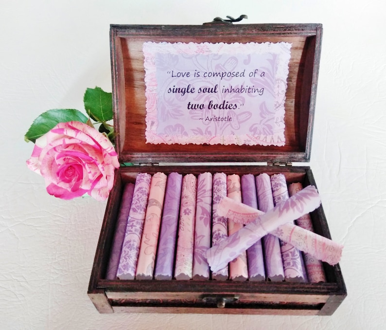 May include: A wooden box with a quote about love by Aristotle inside. The quote reads "Love is composed of a single soul inhabiting two bodies." The box is open and contains a stack of small, rolled-up pieces of paper with a floral pattern.