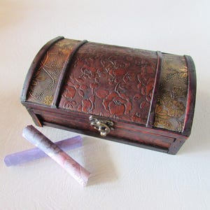 May include: A wooden treasure chest with a floral embossed design and gold accents. The chest is closed and has a metal latch. Two rolled up pieces of paper are in front of the chest.