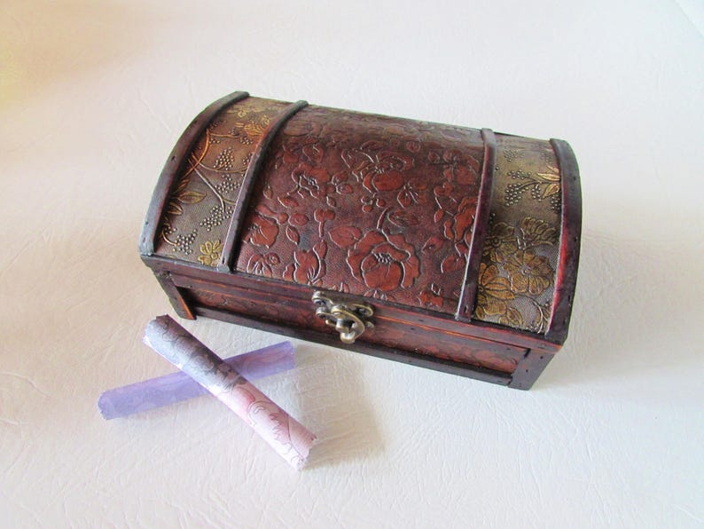 May include: A wooden treasure chest with a floral embossed design and gold accents. The chest is closed and has a metal latch. Two rolled up pieces of paper are in front of the chest.