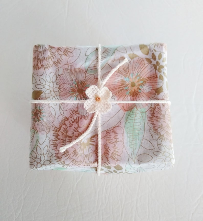 May include: A gift wrapped in white paper with a floral pattern. The paper is tied with white twine and a small white crocheted flower with a brown bead in the center.