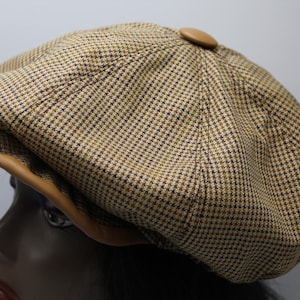 May include: A brown and tan plaid newsboy cap with a leather brim and a button on top. The cap is made of a woven fabric with a small checkered pattern. The brim is a light brown color.