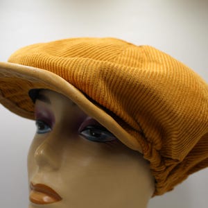 May include: Mustard yellow corduroy newsboy cap with a rounded crown and a stiff, curved brim. The hat is displayed on a mannequin head, showcasing its shape and texture. The cap's color is a warm, earthy tone.