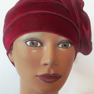 May include: A burgundy velvet hat with horizontal stitched detailing. The hat has a draped design on the side. The hat is shown on a mannequin head with brown eyes and dark red lipstick.