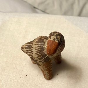May include: A small, handcrafted ceramic duck figurine. The duck has a brown and cream-colored body with a reddish-brown beak. The figurine is standing on four small legs.