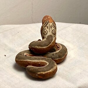May include: A hand-painted, coiled snake figurine. The snake is primarily brown with white and gray stripes, and a detailed head with white and black markings. The figurine is resting on a white surface.