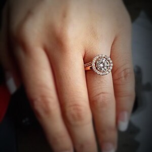 May include: A close-up of a hand wearing a gold engagement ring with a large, round, clear diamond surrounded by smaller diamonds.