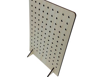 May include: A light-colored wooden pegboard with numerous evenly spaced holes. The board is rectangular with rounded corners and is supported by two angled feet. The pegboard is designed to hold small items.