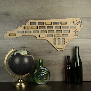 May include: A wooden wall decor shaped like the state of North Carolina with slots to hold wine corks. The decor is mounted on a wood-grain wall. There is a globe, a green glass bottle, and two beer bottles in the foreground.