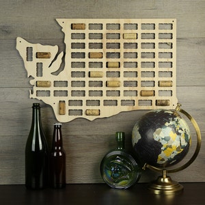 May include: Wooden wall decor shaped like the state of Washington with cork slots for holding wine bottle corks. The decor is on a wood background with two bottles of wine and a globe.
