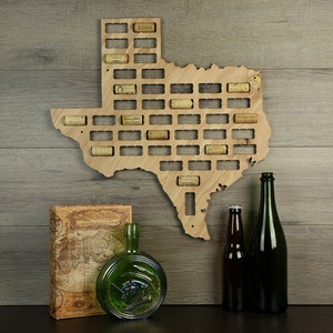 May include: A wooden cutout of the state of Texas with cork slots for wine bottle stoppers. The cutout is mounted on a wood panel background. Two glass bottles are in the foreground.