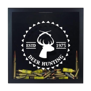 May include: A square black shadow box frame containing a collection of various metallic bullet casings and shotgun shells at the bottom. The glass front features a white graphic of a deer head, crossed rifles, and the text ESTD 1975 DEER HUNTING.