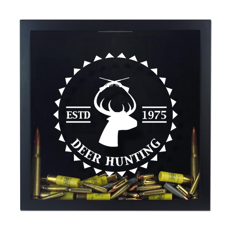 May include: A square black shadow box frame containing a collection of various metallic bullet casings and shotgun shells at the bottom. The glass front features a white graphic of a deer head, crossed rifles, and the text ESTD 1975 DEER HUNTING.