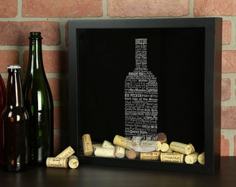 May include: A black shadow box with a wine bottle silhouette made of words related to wine. The bottle is surrounded by cork stoppers. The shadow box is sitting on a brick surface.