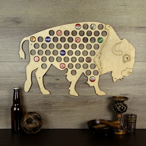 May include: Wooden wall-mounted bottle cap holder shaped like a bison. The holder has 100 holes for bottle caps. The bison is made of light brown wood and is standing on a wooden surface.