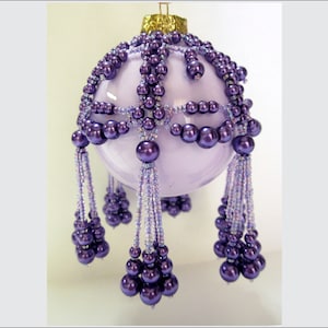Pode incluir: Um enfeite de Natal de vidro roxo com um design de contas e uma borla roxa. O enfeite é decorado com pequenas contas roxas e contas roxas maiores.