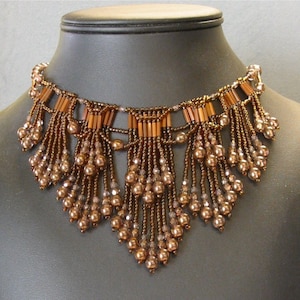 Peut inclure: Un collier de perles brun avec un design en frange. Le collier est fait de petites perles brunes et de plus grosses perles brunes qui créent un effet en cascade.