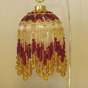 Könnte beinhalten: Ein Weihnachtsbaumschmuck aus Perlen in Gold- und Rottönen. Der Schmuck ist rund und hat einen Perlenbesatz. Er hängt an einem goldenen Ständer.