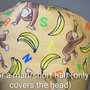May include: A tan scrub cap featuring a repeating pattern of brown monkeys and yellow bananas. The fabric is printed with the letters B, A, N, A, N, A in various colors including red, green, blue, and yellow scattered across the surface.