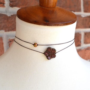 May include: A double-strand choker necklace featuring a thin dark cord. The lower strand displays a dark brown, four-petaled flower-shaped charm, while the upper strand is accented with a single, small, iridescent copper-colored bead.