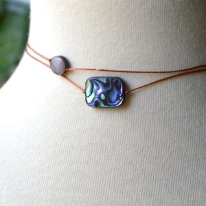 May include: Abalone shell choker necklace. The necklace features a rectangular abalone shell pendant with iridescent blue and green hues. The pendant is strung on a thin, brown cord, with a small, round bead on the side.