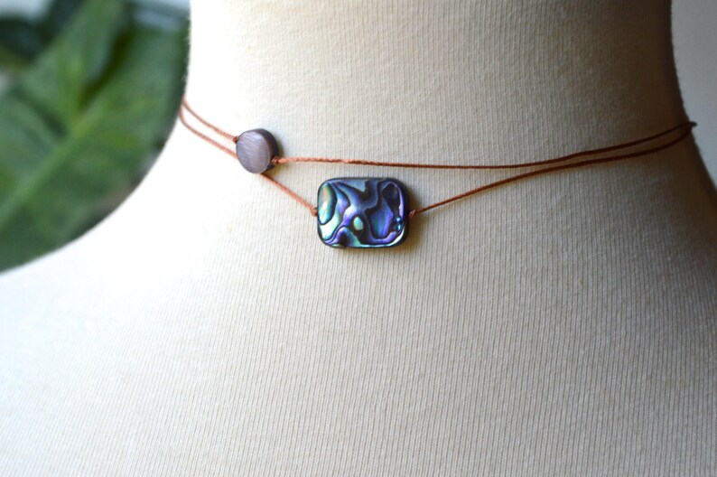 May include: Abalone shell choker necklace. The necklace features a rectangular abalone shell pendant with iridescent blue and green hues. The pendant is strung on a thin, brown cord, with a small, round bead on the side.