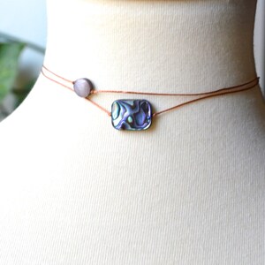 May include: Abalone shell choker necklace. The necklace features a rectangular abalone shell pendant with iridescent colors of blue, green, and purple. A small, round, gray bead is on the side. The necklace is displayed on a mannequin.